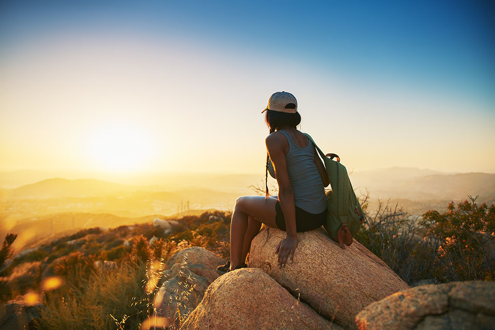Woman hiker sitting on rock on top of hill while looking at sunset over San Diego California
